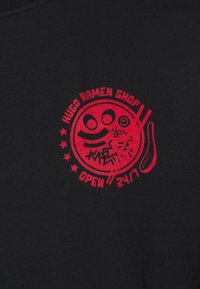 T-shirt en coton noir avec un graphisme circulaire rouge comportant "Hugo Ramen Shop," un visage souriant, des étoiles et "Ouvert 24/7."