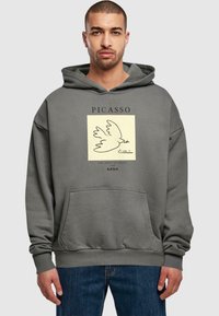 Mikina s kapucí v tlumené šedé látce, která obsahuje grafický potisk holubice uvnitř čtverce, s nápisem "PICASSO" nahoru a "HOLUBICE MÍRU" dole.