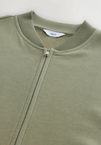 Sudadera verde claro con cremallera y cuello acanalado. La tela principal es suave con un ligero brillo, acentuada por la cremallera metálica.