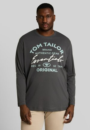 Man die een donkergrijs shirt met lange mouwen draagt met blauwe en witte "Tom Tailor Essentials Original" tekst en lichtbeige broek.