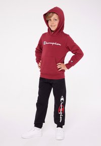 Felpa rossa con cappuccio e logo bianco "Champion", abbinata a pantaloni neri con un logo sul lato e sneakers bianche. Tessuto morbido, vestibilità comoda.