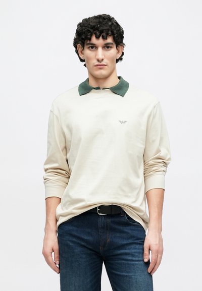 Bēša garroku polo krekls ar tumši zaļu apkakli, ar nelielu logo krūtīs, kombinēts ar tumši ziliem džinsiem.