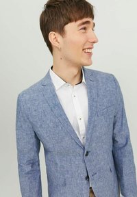 Jasnoniebieska marynarka z teksturowanym wykończeniem, zapinana na jeden guzik, z dwoma przednimi kieszeniami, noszona na białej koszuli z subtelnym kołnierzykiem.