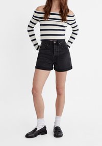 Mujer con blusa blanca de rayas azul marino y hombros descubiertos, shorts de mezclilla negros de tiro alto, calcetas blancas y mocasines negros, de pie con las manos en la cintura.