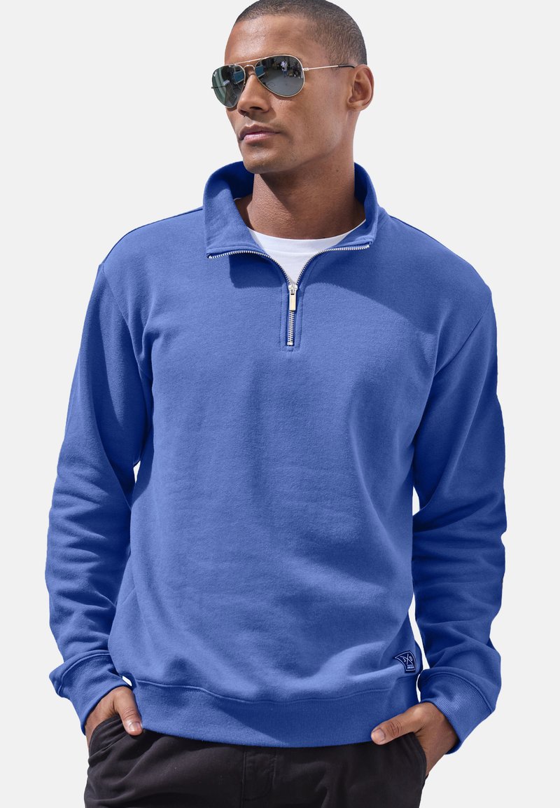 Blue zip-up sweatshirt with a high collar, soft texture, ribbed cuffs, and a small logo accent near the hem. Worn over a white shirt.