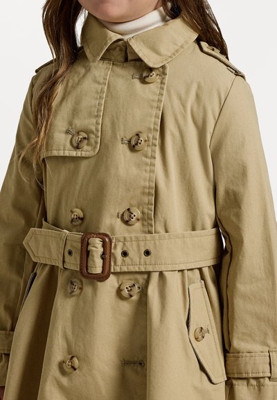 Tan bomullstrenchcoat med dubbelknäppt design, stora knappar, bälte och axelpass. Har raka linjer och sidofickor.