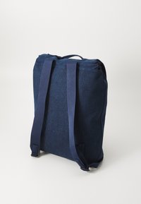 Temno moder jeans nahrbtnik z dvema naramnicama, zgornjim ročajem in trdno zasnovo. Gladka tekstura brez vidnih kovinskih podrobnosti.