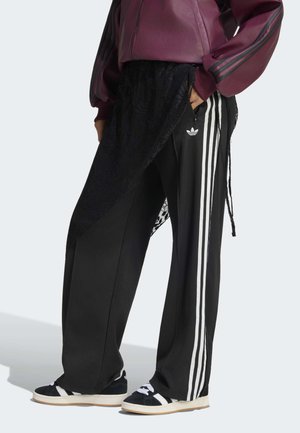 Person iført sorte Adidas træningsbukser med hvide striber, sort blondeoverlay, sort-hvide sneakers og en vinrød jakke med sorte striber.