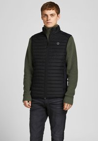 Sort kviltet vest med høj krave, parret med en grøn ribbet sweater. Vesten har en lynlåslukning foran og et logo på brystet.