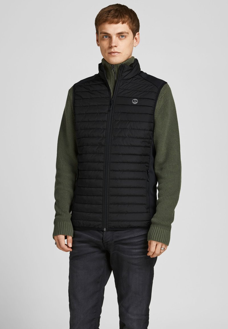 Sort kviltet vest med høj krave, parret med en grøn ribbet sweater. Vesten har en lynlåslukning foran og et logo på brystet.