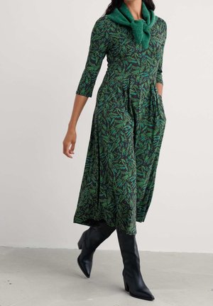 Frau in einem grünen, gemusterten Midikleid mit Dreiviertelärmeln und schwarzen Stiefel mit Absatz, die einen grünen Pullover über die Schultern gelegt trägt.