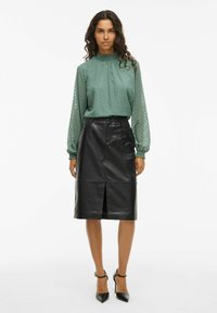 Blusa verde a trama con collo alto, abbinata a una gonna a tubino in pelle nera con un'apertura frontale. Tacchi a spillo neri completano l'outfit.