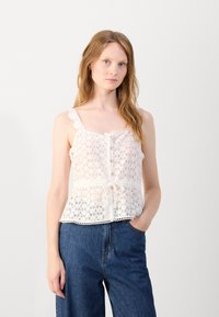 White lace blouse with a V-neck, ruffled straps, button front, drawstring waist, and intricate floral patterns. Paired with blue denim pants.