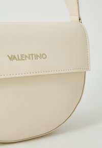 Valentino veske i kremfarget kunstlær med en strukturert, avrundet form. Den har gulllogo, sydd detaljer og justerbar stropp.
