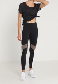 Top atlético negro con detalle de nudo, combinado con leggings negros que presentan acentos en bloque de color en blanco y marrón. Calzado: zapatillas blancas.