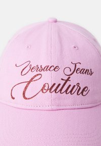 Rozā beisbola cepure, kas izgatavota no teksturētas auduma, ar spīdīgu, uzrakstītu brūnu burtu, kas lasāms "Versace Jeans Couture."
