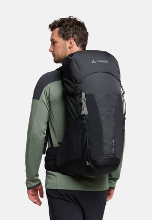 Zaino nero con più scomparti, spallacci imbottiti, un coperchio superiore e una superficie testurizzata. Indossato sulla schiena, abbinato a una maglietta verde.