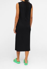 Robe noire sans manches à texture côtelée, de longueur midi avec des fentes sur les côtés, associée à des sandales vertes.