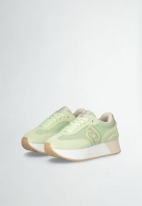 Sneakers verde chiaro realizzati con materiali in rete e suede, dotati di una suola geometrica bianca e branding discreto. Lacci e imbottitura visibili.