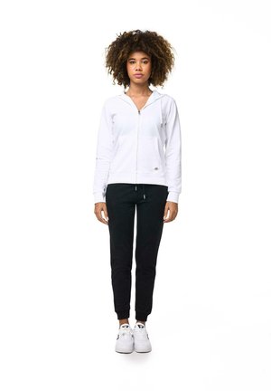 Una mujer de pelo rizado está de pie sobre un fondo blanco, vestida con una sudadera blanca con capucha y cremallera, pantalones negros y zapatillas deportivas blancas.