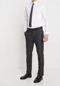 Uomo con camicia bianca, cravatta a righe, pantaloni grigio scuro, cintura marrone scuro e scarpe eleganti marrone scuro, in piedi con le mani nelle tasche.