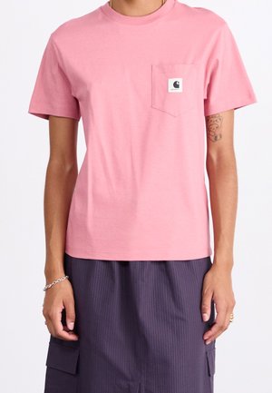 Osoba ubrana w różowy t-shirt Carhartt z kieszenią oraz ciemne spodnie cargo w cienkie prążki, z widocznym tatuażem misia na lewej ręce.