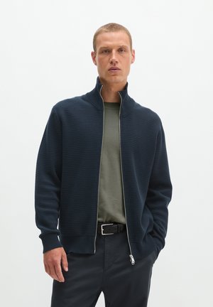 Homme portant un pull zippé bleu foncé sur une chemise vert olive et un pantalon foncé, debout avec une main dans la poche devant un fond uni.