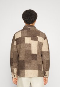 Person set bagfra iført en brun og beige ternet uldjakke med lange ærmer og lyse bukser.