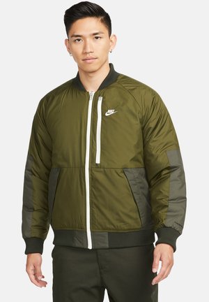 Veste bomber vert olive avec une fermeture éclair sur le devant, des poignets côtelés et le logo blanc de Nike. Elle présente un design de panneaux contrastés et une texture lisse.