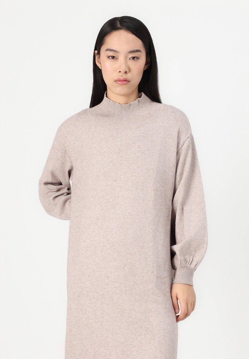 Jasno beżowa sukienka swetrowa z wysokim, falbaniastym dekoltem i długimi rękawami. Wykonana z miękkiego, teksturowanego materiału dzianinowego.