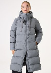 Abrigo acolchado gris con cuello alto, diseño acolchado, cierre de cremallera y detalles de tela con lazos. Mangas largas y largo hasta la mitad de la pantorrilla.