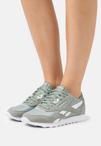 Par de zapatillas Reebok Classic de color verde claro y blanco llevadas en los pies sobre un fondo blanco liso.