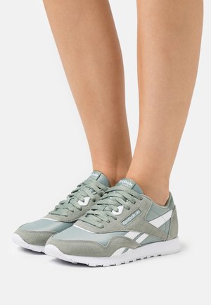 Pár světle zelených a bílých tenisek Reebok Classic na nohou na jednoduchém bílém pozadí.