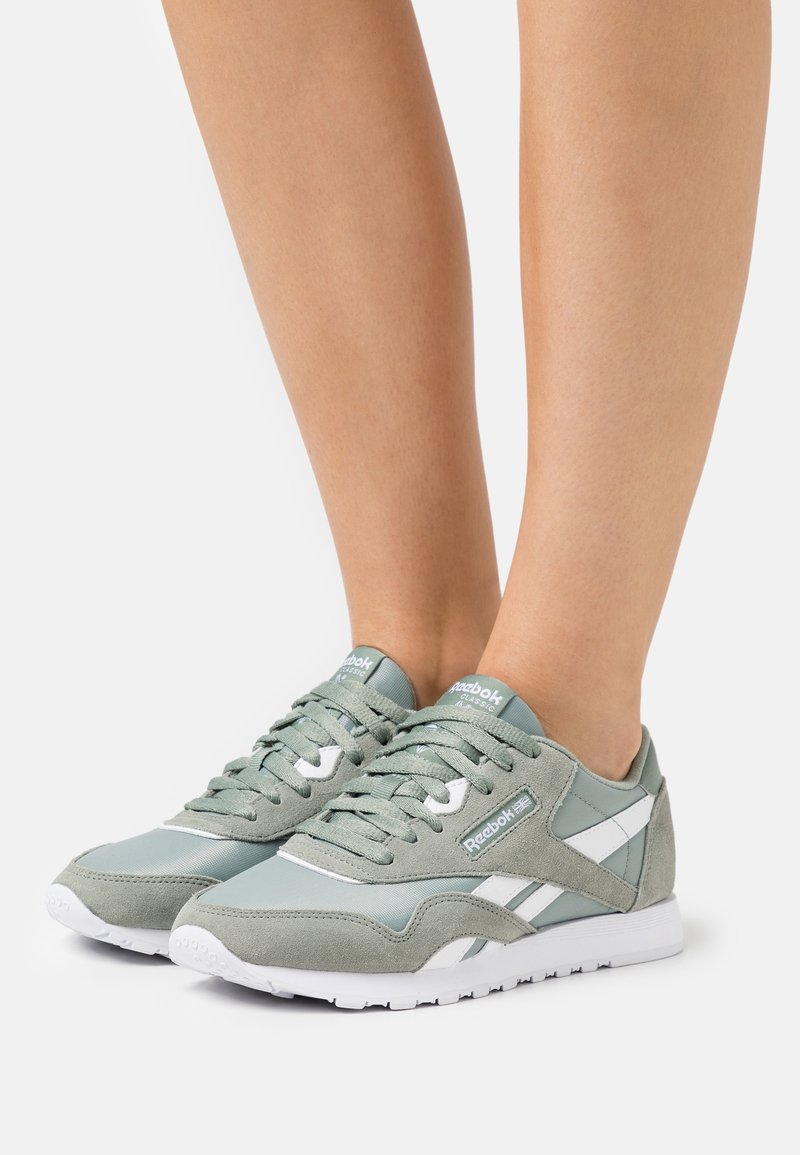 Par de zapatillas Reebok Classic de color verde claro y blanco llevadas en los pies sobre un fondo blanco liso.