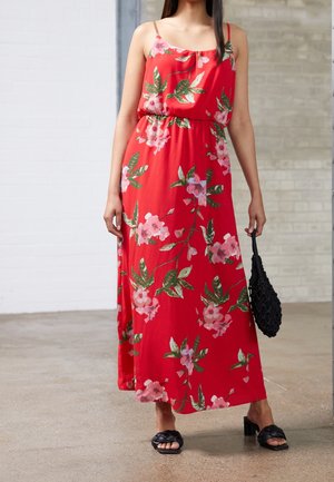 Frau trägt ein rotes, geblümtes Maxikleid mit dünnen Trägern, hält eine schwarze geflochtene Handtasche und trägt schwarze High-Heel-Sandalen mit geflochtenen Riemen.