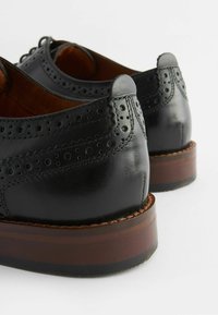 Zapatos de cuero negro con detalles de brogue, presentando una textura suave y un tacón de madera apilado bajo. Realzados con diseños perforados.