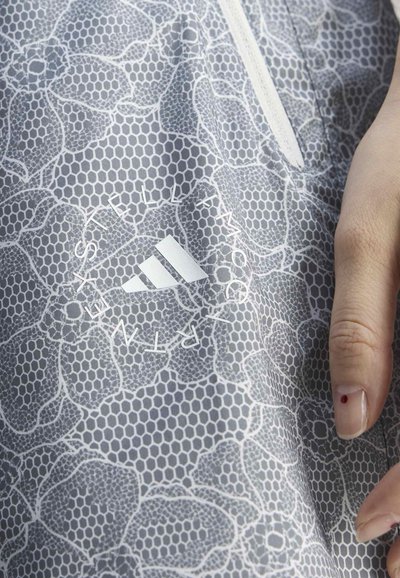 Main reposant sur une veste de sport grise avec motif floral hexagonal, logo Adidas blanc et texte « Stella McCartney » près d'une fermeture éclair blanche.