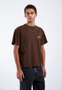 Brown cotton t-shirt with a relaxed fit, short sleeves, and a white embroidered logo on the left chest. Paired with dark denim jeans.
