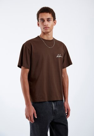 Braunes Baumwoll-T-Shirt mit lockerem Schnitt, kurzen Ärmeln und einem weißen gestickten Logo auf der linken Brust. Kombiniert mit einer dunklen Jeans.