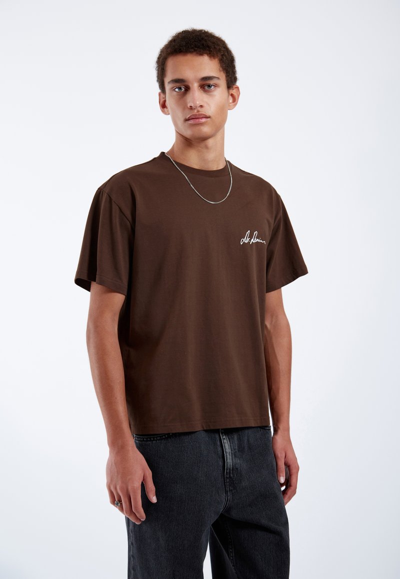Brown cotton t-shirt with a relaxed fit, short sleeves, and a white embroidered logo on the left chest. Paired with dark denim jeans.
