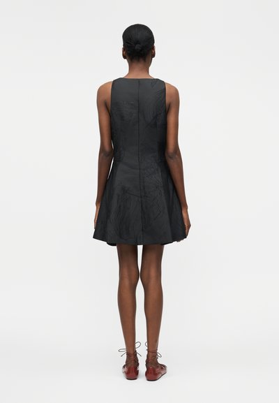 Γυναίκα με σκούρο δέρμα και μαλλιά σε κότσο, που φοράει μαύρο αμάνικο φόρεμα και κόκκινα παπούτσια με κορδόνια, στέκεται πλάτη σε λευκό φόντο.