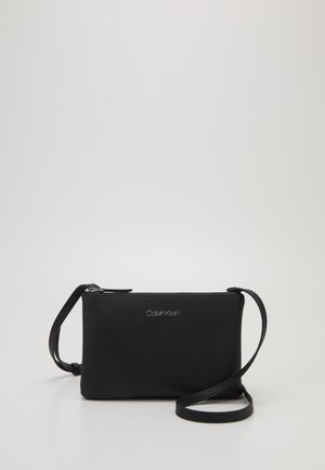 Czarna skórzana torebka na ramię Calvin Klein z zamkiem błyskawicznym i regulowanym paskiem na jednolitym jasnym tle.