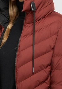 Chaqueta acolchada roja con un patrón en chevrón, cremallera completa y acentos en negro. Presenta una capucha ajustable con cordón y tope de plástico.