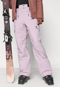 Lyse lilla skibukser med høj talje, sidelommer og lynlåsdetaljer, parret med sorte skistøvler og et ski.