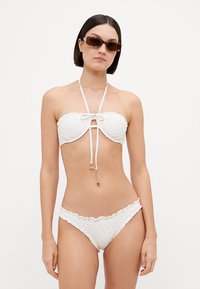 Ensemble de bikini blanc comprenant un haut sans bretelles avec des détails en dentelle et un nœud, associé à un bas de bikini à bord festonné. Texture lisse.