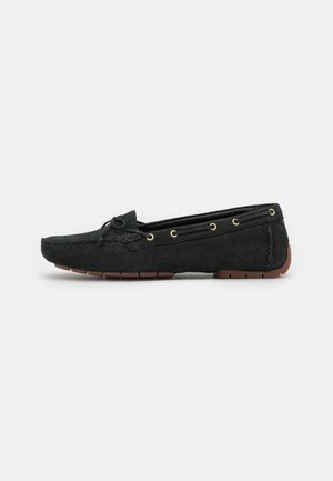Zwarte suède mocassin loafer met bruine rubberen zool, decoratieve strik en gouden oogjes langs de zijkanten.