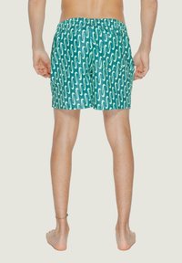 Costumi da bagno verdi con un motivo ondulato bianco, vita elasticizzata e tasche laterali, realizzati in tessuto leggero, visti da dietro.