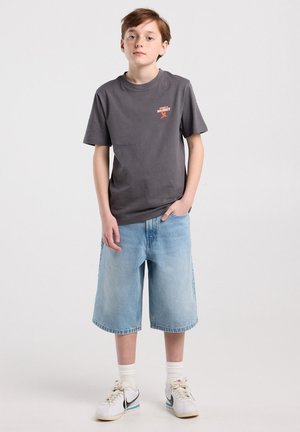 Junge trägt ein dunkelgraues T-Shirt, hellblaue Jeans-Shorts, weiße Socken und weiße Turnschuhe mit schwarzem Swoosh, steht mit einer Hand in der Tasche.