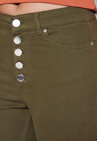 Jean taille haute vert olive avec cinq boutons décoratifs en argent, poches avant et coutures contrastantes. Texture lisse dans l'ensemble.