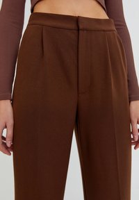 Pantalons marron ajustés avec une taille structurée et des plis, affichant une texture lisse. Probablement fabriqués dans un tissu doux et extensible.